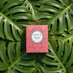 Bougie parfumée Tahiti Candles Tropical Fruit dans son emballage rose à motifs polynésiens sur des feuilles de monstera
